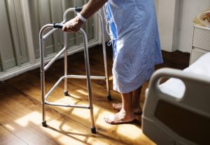 Elderly man in lightblue hospital gown walking with walker away from his hospital bed in the morning.