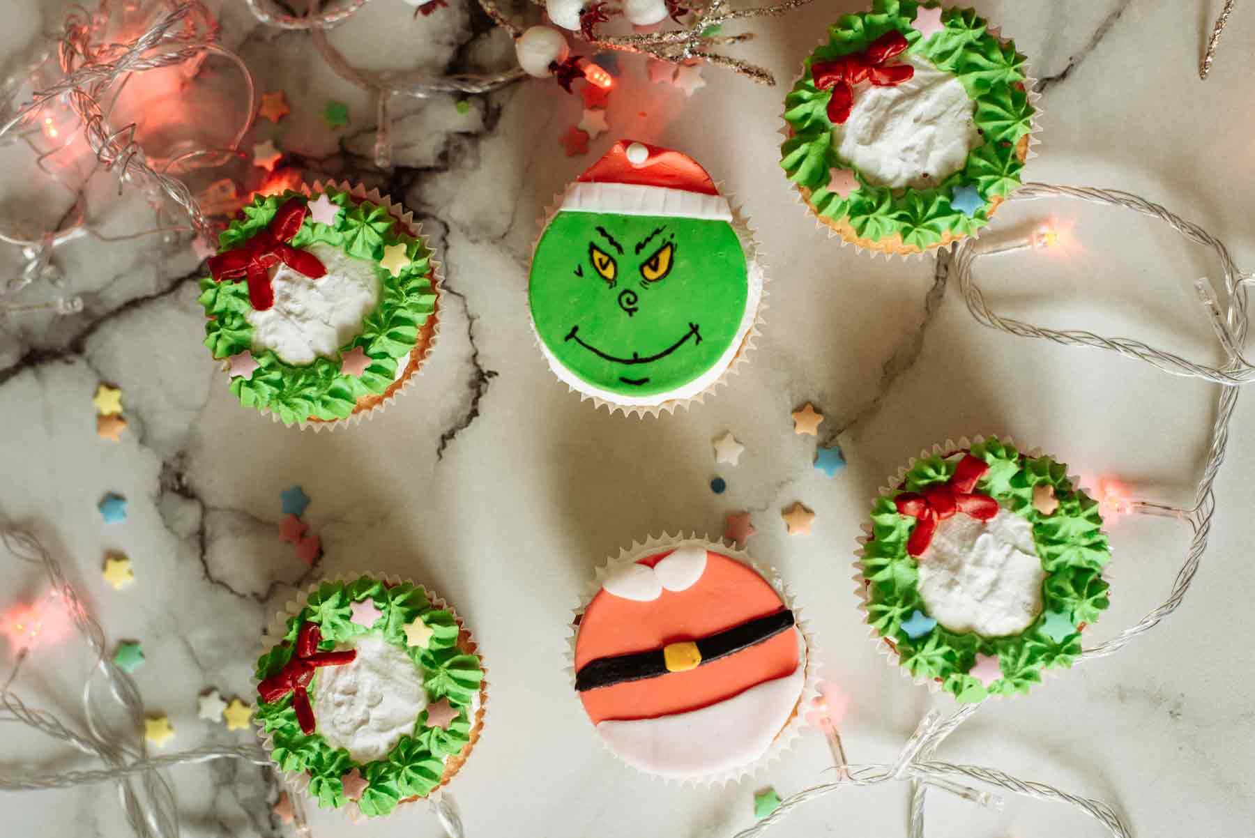 Six Christmas cupcakes (one decorated with a grinch face) with Christmas lights and marzipan stars scattered on a white surface