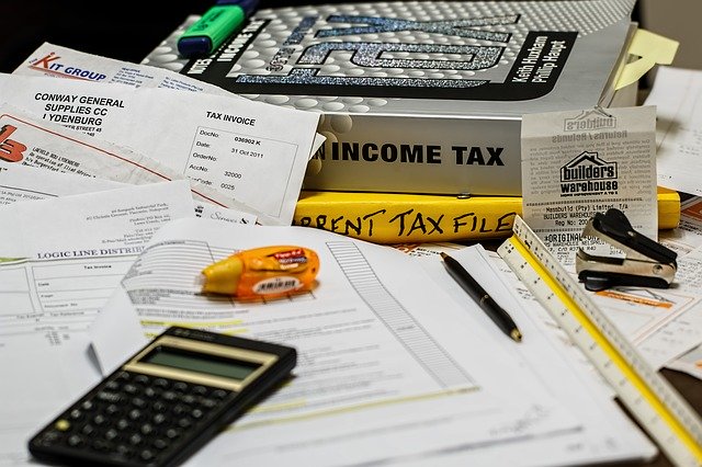 Image of filing income tax, with papers strewn across a desk with a calculator, green highlighter, white-out pen and thick silver income-tax book