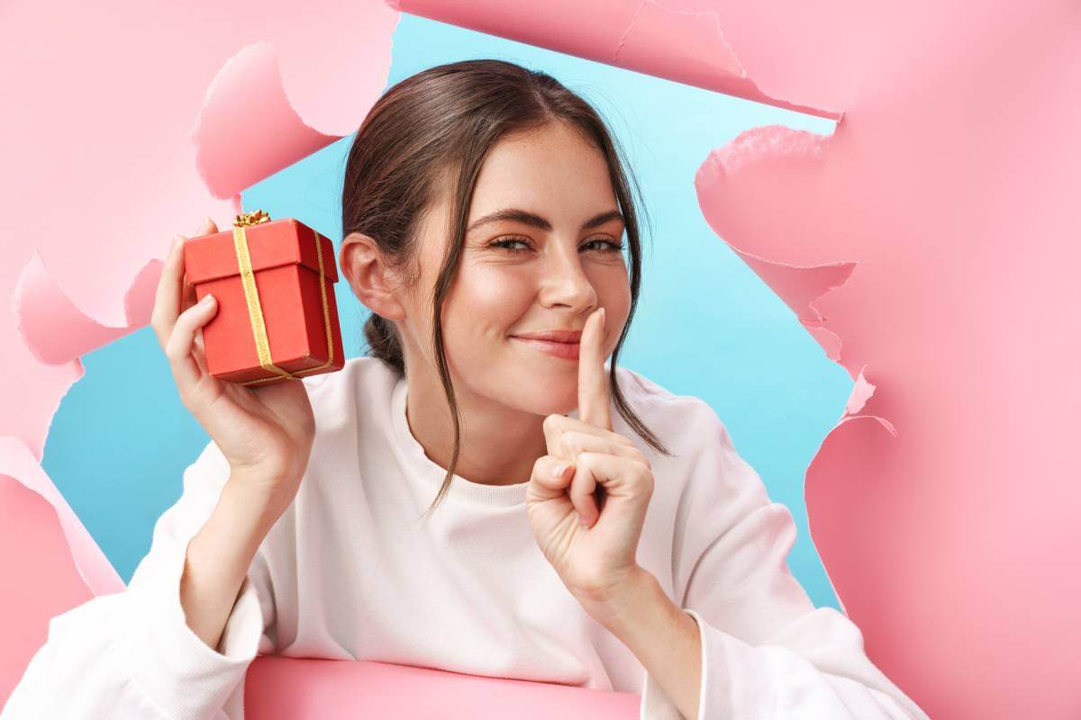 Young girl in white sweater holds a wrapped gift with fingers on lips on blue and pink background.