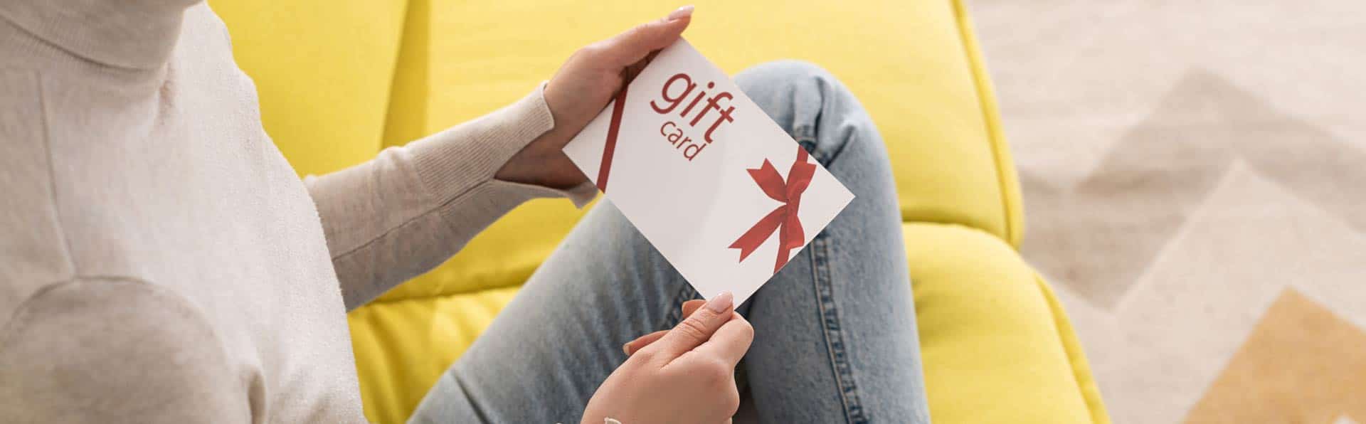 Woman in blue jeans sits on yellow couch and holds a gift card envelope
